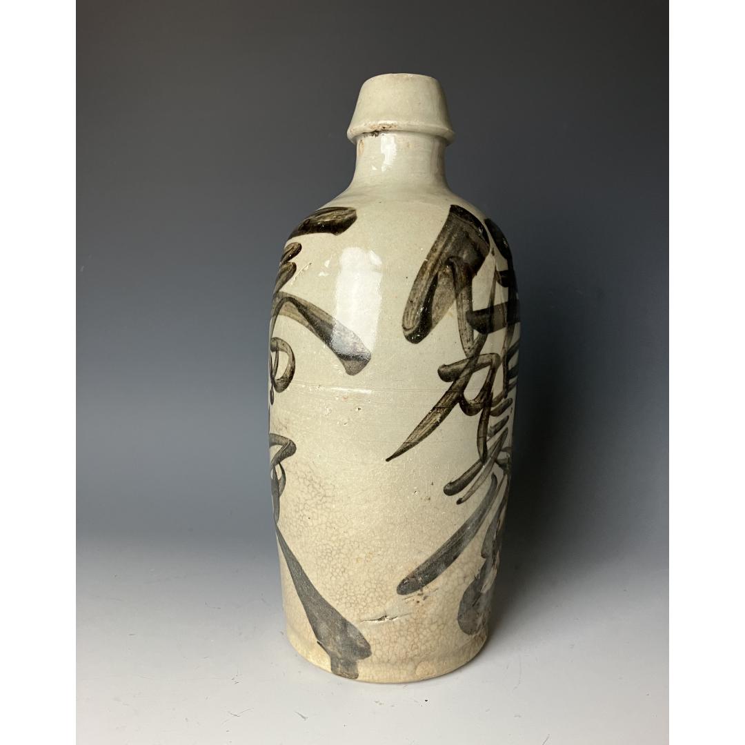 An angled view of a vintage Japanese Tokkuri sake bottle, displaying bold kanji characters on a textured ceramic surface with a visible crackle glaze effect.