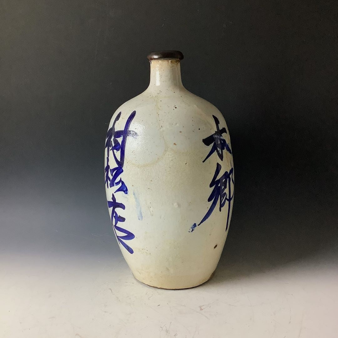 A tall Japanese Tokkuri sake bottle displaying blue calligraphy, with a crackled glaze and a brown neck, set against a dark background.