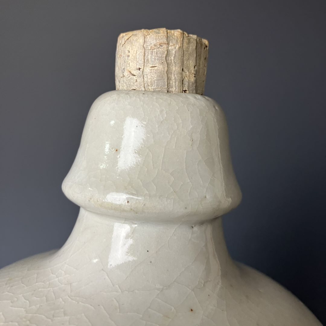 Close-up of the top of a ceramic sake bottle with a crackled glaze finish and a rustic, textured cork stopper, set against a muted grey background.