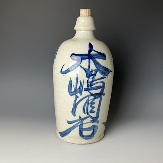 A Japanese ceramic sake bottle (tokkuri) with blue calligraphy on a white crackled glaze background, featuring a cork stopper, against a grey backdrop.