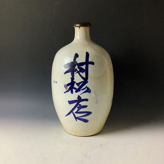 A traditional Japanese Tokkuri sake bottle with dark blue calligraphy on a white glaze, featuring a brown rim, presented against a black background.