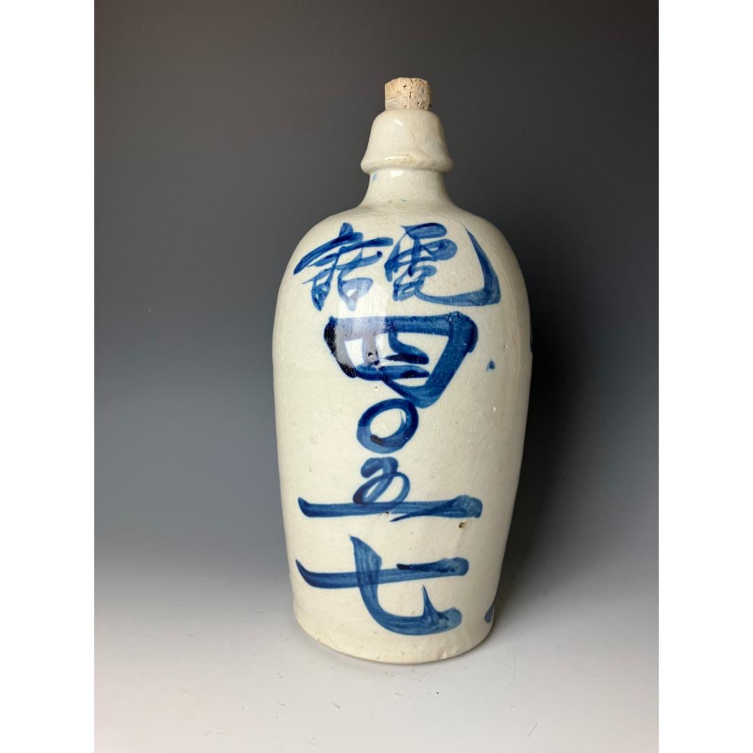A ceramic sake bottle with blue kanji script, cork stopper, and a white crackle glaze, set against a neutral background.