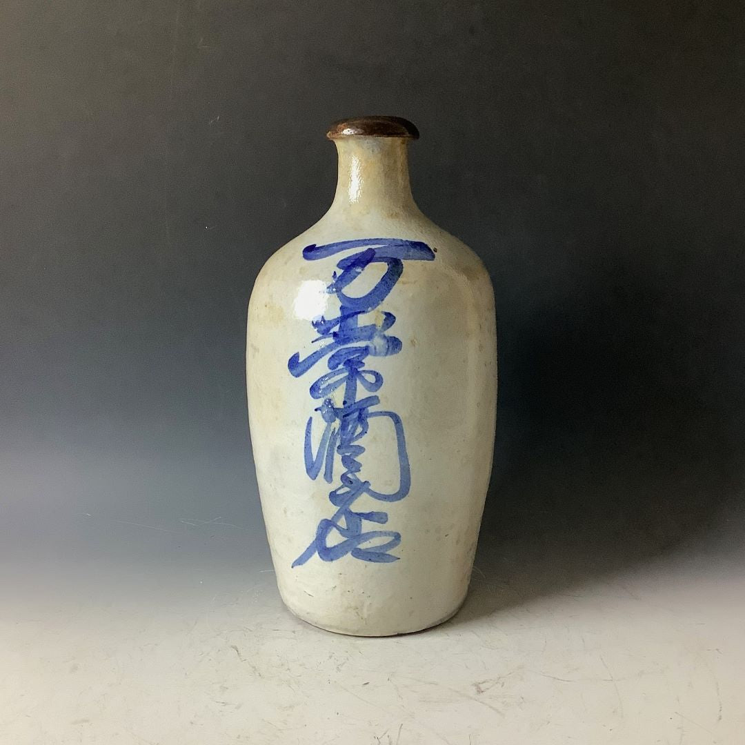 A traditional Japanese tokkuri (sake bottle) viewed from a different angle, displaying a series of blue calligraphic characters on its speckled beige surface. The narrow neck of the bottle shows a dark brown rim, and it stands against a muted gray backdrop which highlights the bottle's form and the striking blue script.