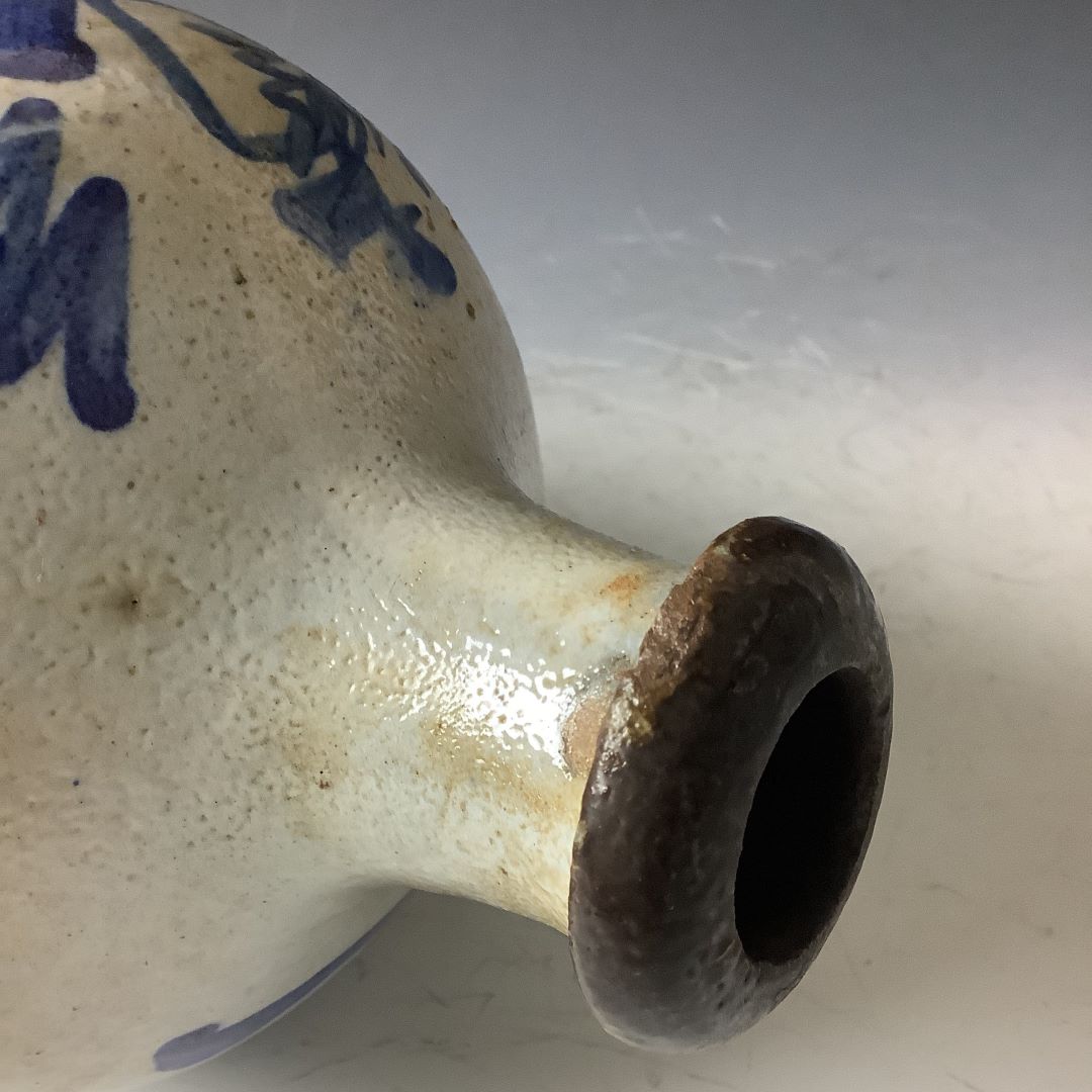 A close-up of the top section of a traditional Japanese tokkuri (sake bottle), highlighting the textured beige glaze and the dark brown rim around the opening. Blue calligraphic characters are partially visible, curving around the bottle's shoulder. The image focuses on the craftsmanship of the glaze and the contrast between the beige body and the dark rim.
