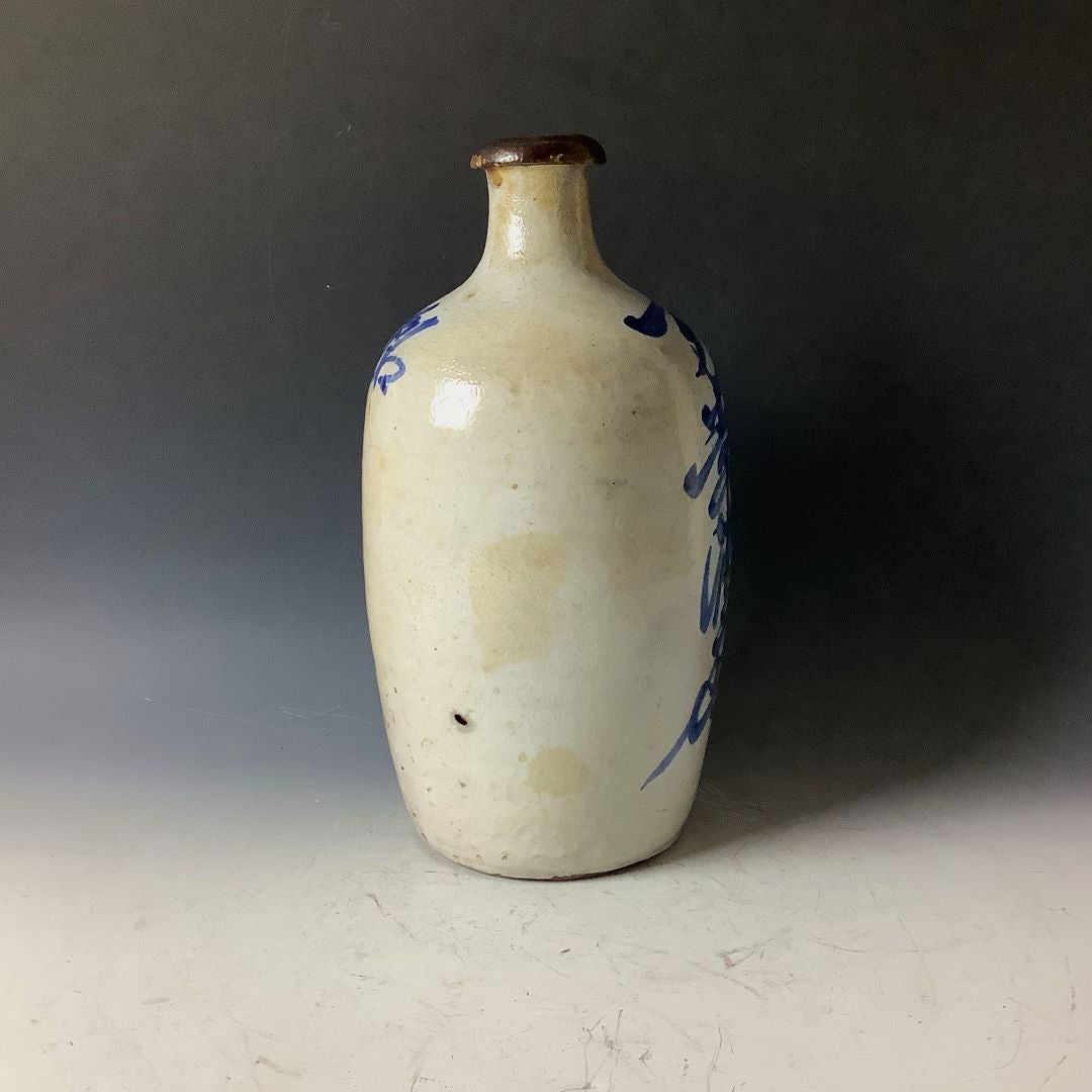 A traditional Japanese tokkuri (sake bottle) with minimal blue brushstroke embellishments on its speckled beige surface, showcasing a simple and elegant design. The bottle features a dark brown rim around the narrow neck, set against a soft gray background that subtly complements the bottle's natural color palette.