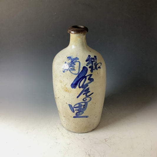  A traditional Japanese tokkuri (sake bottle) with a narrow neck and wider base, featuring calligraphic blue brush stroke characters on a speckled beige background. The bottle's rim is dark brown, possibly from use or a glazing effect. The background is a neutral gray, contrasting with the bottle's light color.