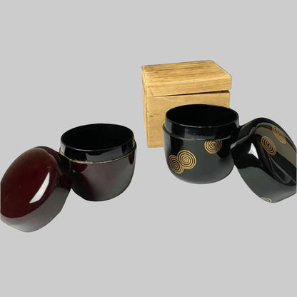 Two open 19th-century Japanese lacquered makie natsume tea caddies, one in deep brown and the other in black with gold spiral designs, accompanied by a wooden box, highlighting their traditional craftsmanship and elegant design.