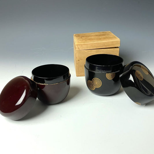 Two open 19th-century Japanese lacquered makie natsume tea caddies, one in deep brown and the other in black with gold spiral designs, accompanied by a wooden box, highlighting their traditional craftsmanship and elegant design.
