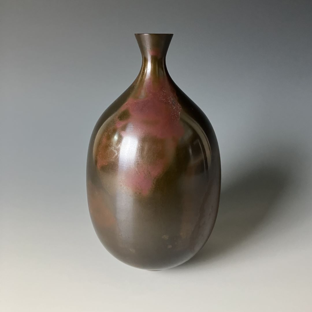 An elegant Japanese copper vase displayed against a soft grey background, with a sleek, rounded body and a narrow neck. The surface reflects a subtle patina that gives the vase a dynamic and organic feel.
