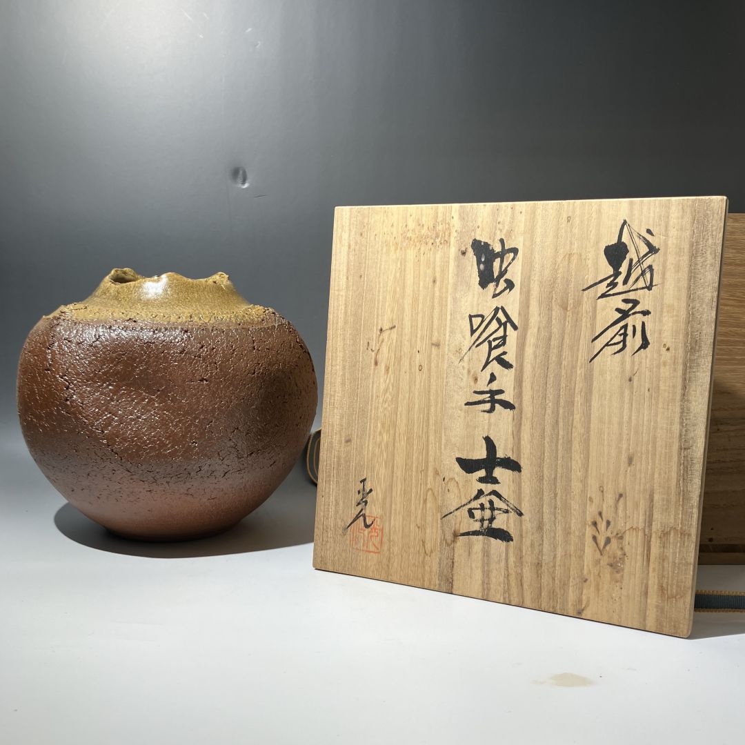 Brown ceramic vase next to a wooden plaque with calligraphy on a gray background