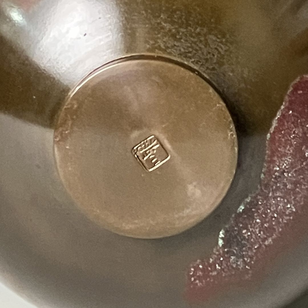  Close-up of the bottom of a copper vase, displaying a maker's mark or signature stamp, indicative of its artisanal Japanese craftsmanship. The texture around the stamp shows the vase's unique patina, blending colors and sheen.