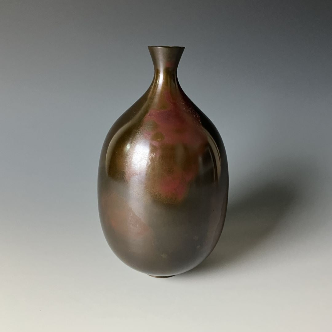  A solitary Japanese copper vase on a grey background, with a smooth, tapered shape and a patina showcasing shades of dark and reddish copper, exemplifying traditional elegance in its minimalist design.