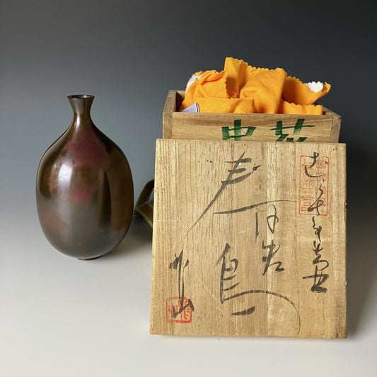 A handcrafted Japanese copper vase with an elegantly tapered neck and a patinated surface is paired with a wooden box adorned with calligraphy and containing orange cloth, evoking a sense of traditional elegance and the art of Ikebana.