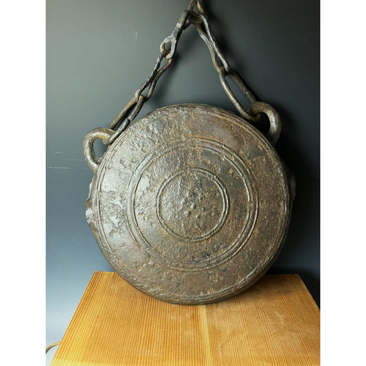 An aged metal Japanese Waniguchi gong with chain, featuring raised designs, displayed on a wooden stand against a dark background.