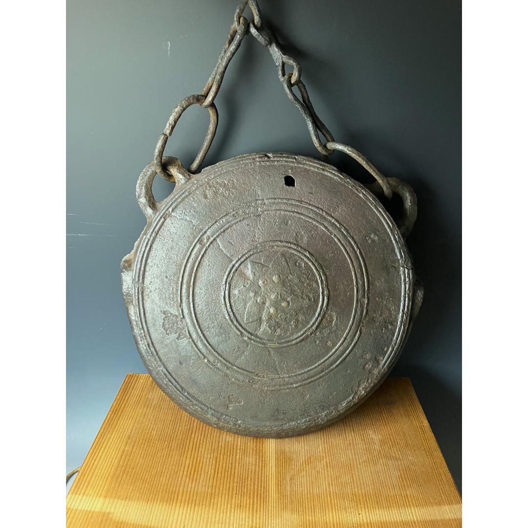 An aged metal Japanese Waniguchi gong with chain, featuring raised designs, displayed on a wooden stand against a dark background.