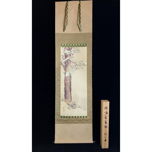  An East Asian hanging scroll depicts a plum tree in bloom with a decorative border, suspended by green cords, accompanied by a wooden box with East Asian script. The background is black.