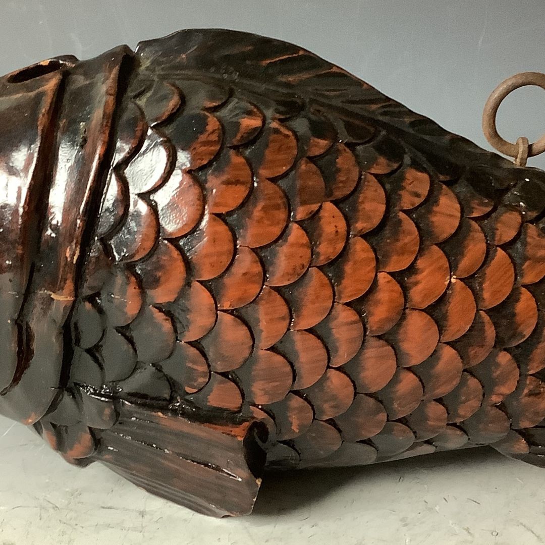 A close-up of the body of a wooden fish sculpture, showcasing its intricately carved and polished scales in a rich, dark brown hue with a lustrous sheen, accompanied by a metal ring for hanging, against a soft gray background.