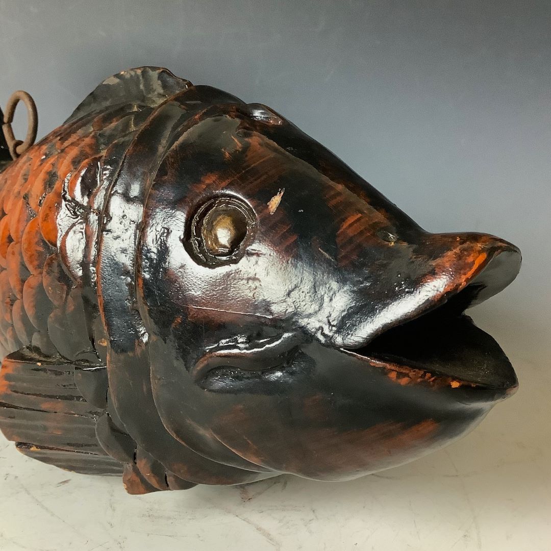 A close-up of a sculpted wooden fish head with a dark, glossy finish, featuring detailed eyes, mouth, and scale texture, with a metal ring attached above, set against a gradient gray backdrop.