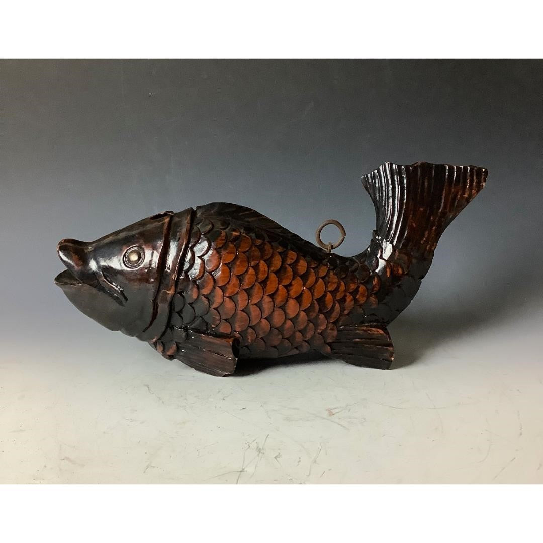 A decorative, dark brown, wooden fish sculpture with detailed scales and fins, featuring a metallic ring on its back for hanging, displayed on a gray surface against a gradient gray background.