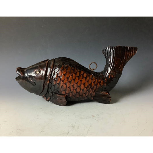 A decorative, dark brown, wooden fish sculpture with detailed scales and fins, featuring a metallic ring on its back for hanging, displayed on a gray surface against a gradient gray background.