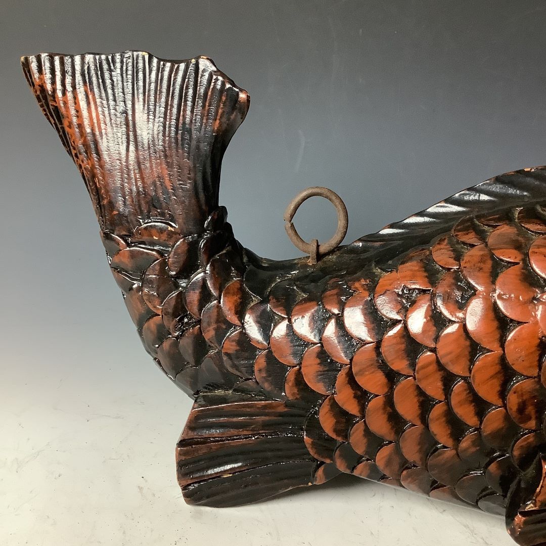 A side view of a wooden fish sculpture with a striking tail fin and detailed scales, finished in dark glossy brown with highlights. A metal hanging ring is attached near the tail fin, against a smooth gradient gray background.
