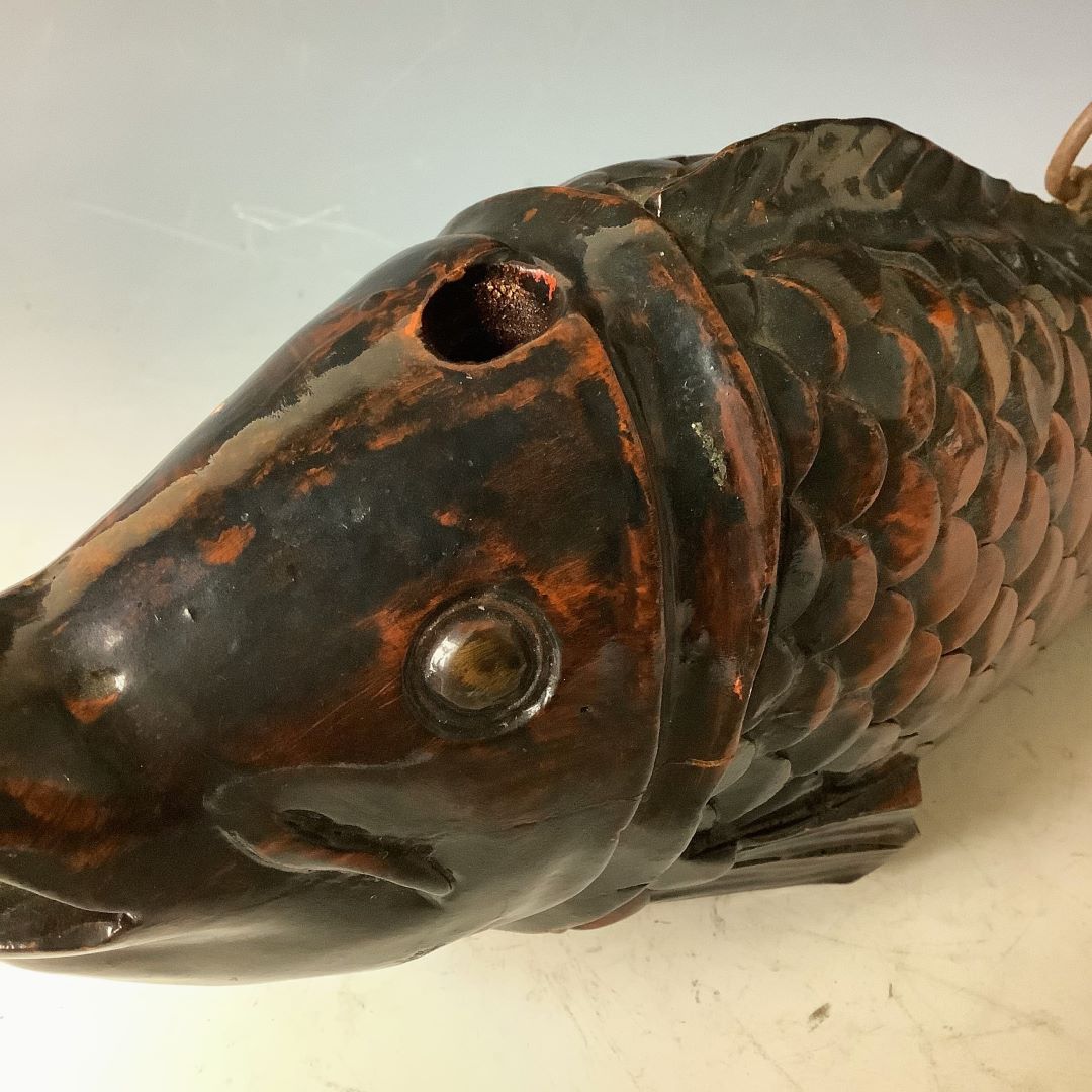 Close-up of the head of a wooden fish sculpture with a shiny, dark patina finish. The fish has a carved eye and mouth, textured scales, and a hole on top where a piece is missing, set against a light gray background with slight scuff marks.