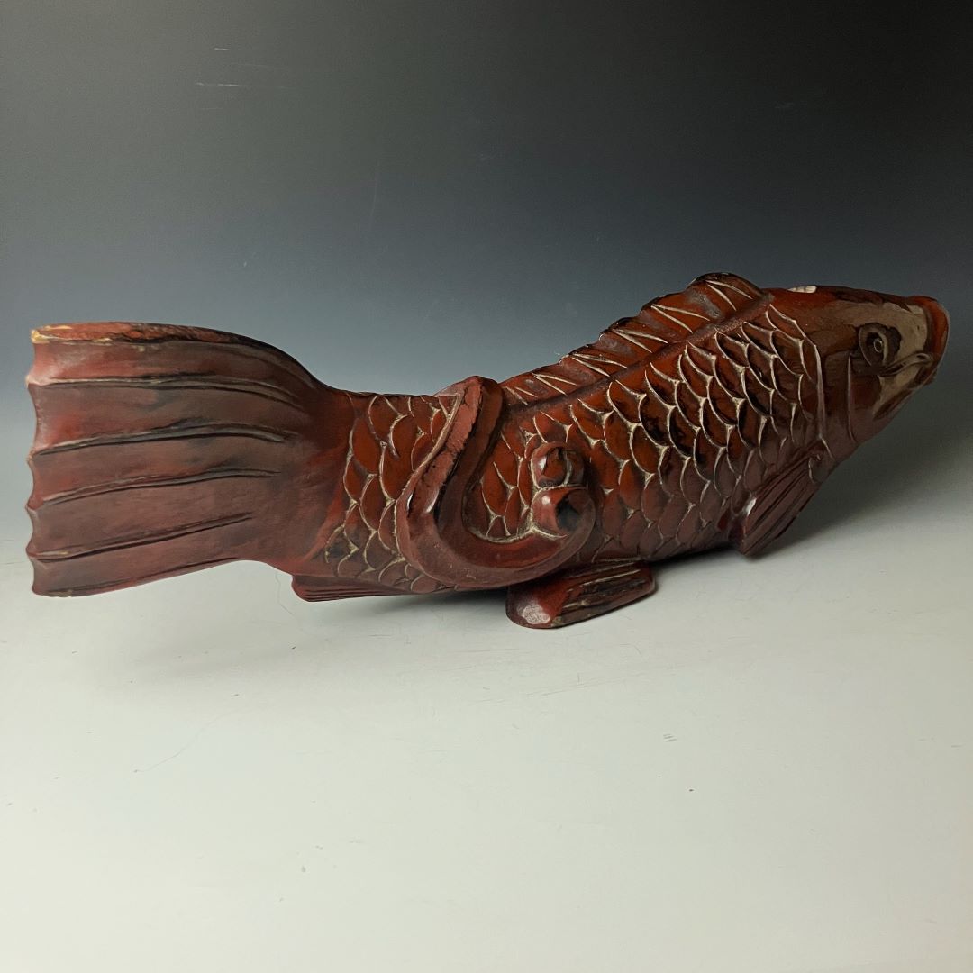  This image displays a full side view of an antique wooden fish carving, with a rich reddish-brown patina, detailed scales, and a gracefully carved tail. The fish's eye is detailed and expressive, adding life to the carving. The object stands on a flat surface against a neutral background, highlighting its artistic qualities and the fine craftsmanship.