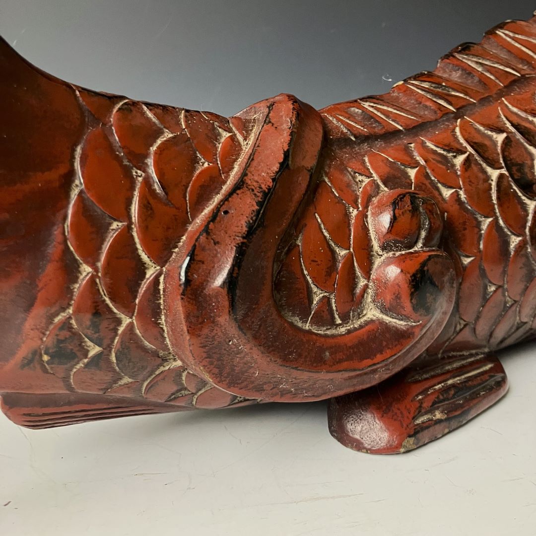 Close-up of the tail section of an antique wooden fish carving, highlighting the detailed scale pattern and the aged red-brown patina with black accents, along with the curved form and the weathered surface that adds to its antique character.