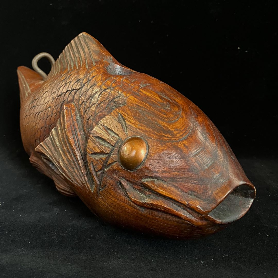 A wooden carving of a fish's head with a prominent eye, textured scales, and detailed fins, featuring a metal ring on the top, set against a black background.