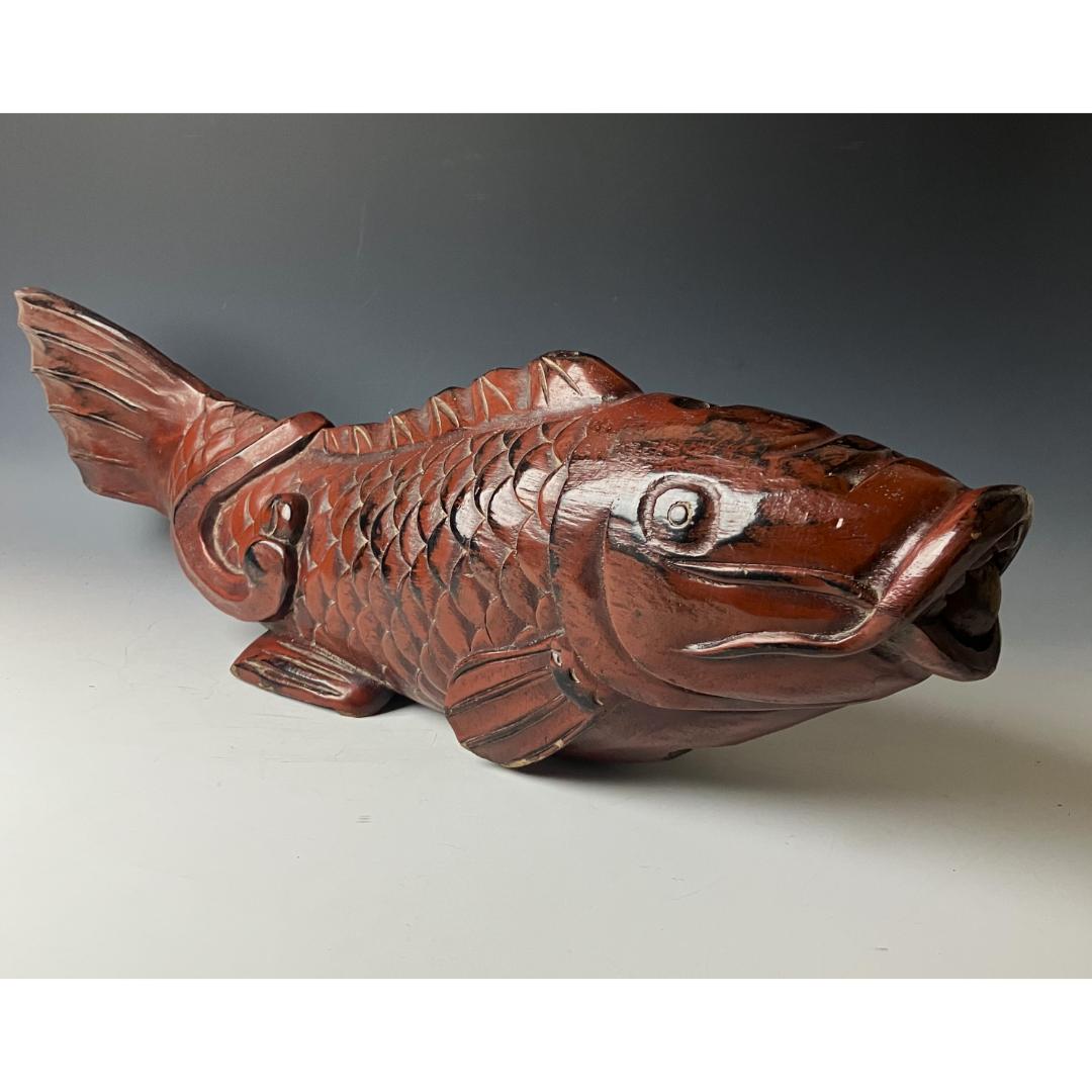 An antique Japanese kitchenware item, designed as a detailed wooden fish carving. The fish is predominantly brown with darker shading to accentuate its scales and features. The fish is positioned with the head and tail bent slightly upwards.