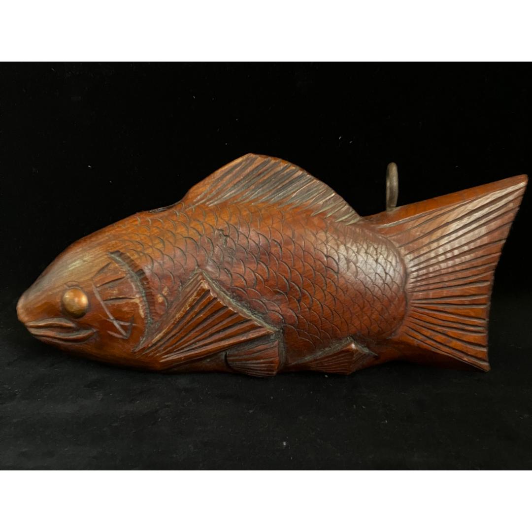 A close-up of a wooden fish sculpture with a metal loop protruding from its back, intricately carved to depict scales, fins, and facial features, presented against a black background.