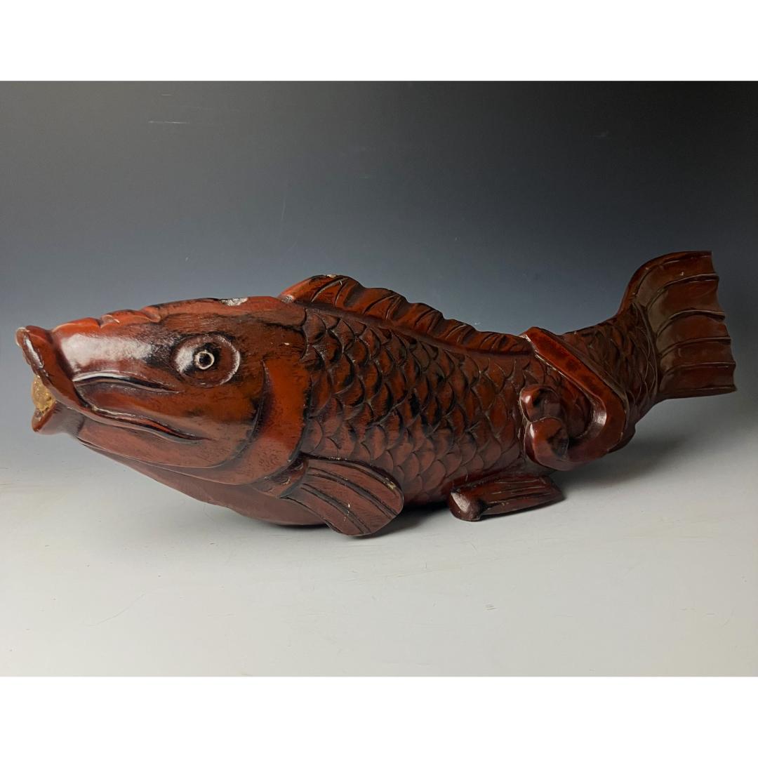 An antique Japanese kitchenware item, designed as a detailed wooden fish carving. The fish is predominantly brown with darker shading to accentuate its scales and features. The fish is positioned with the head and tail bent slightly upwards.