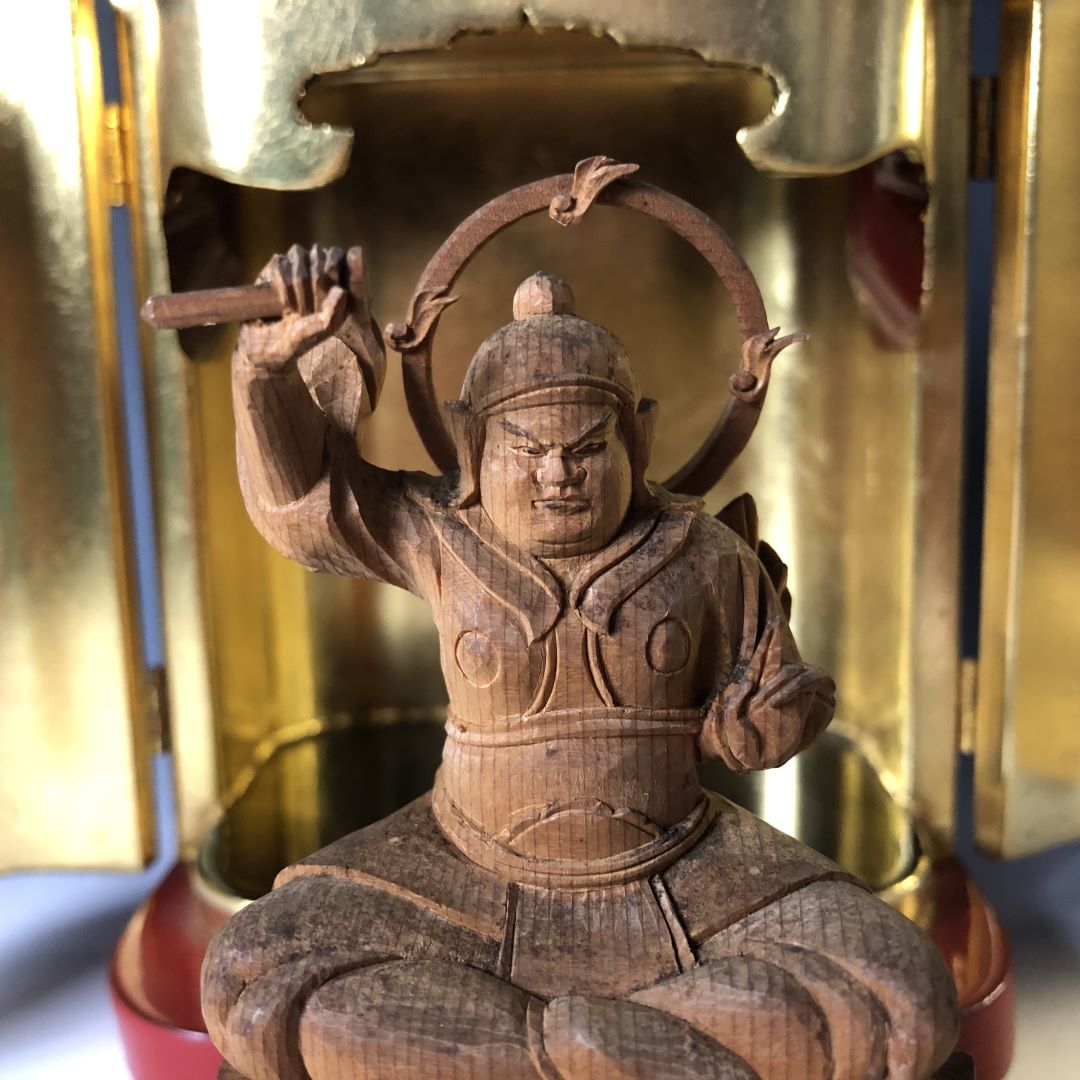 A close-up of a hand-carved wooden Myoken Bosatsu statue within a golden Zushi cabinet. The figure, depicted with a stern expression, is dressed in traditional attire with a helmet and holds aloft a missing sword, framed by a circular halo.