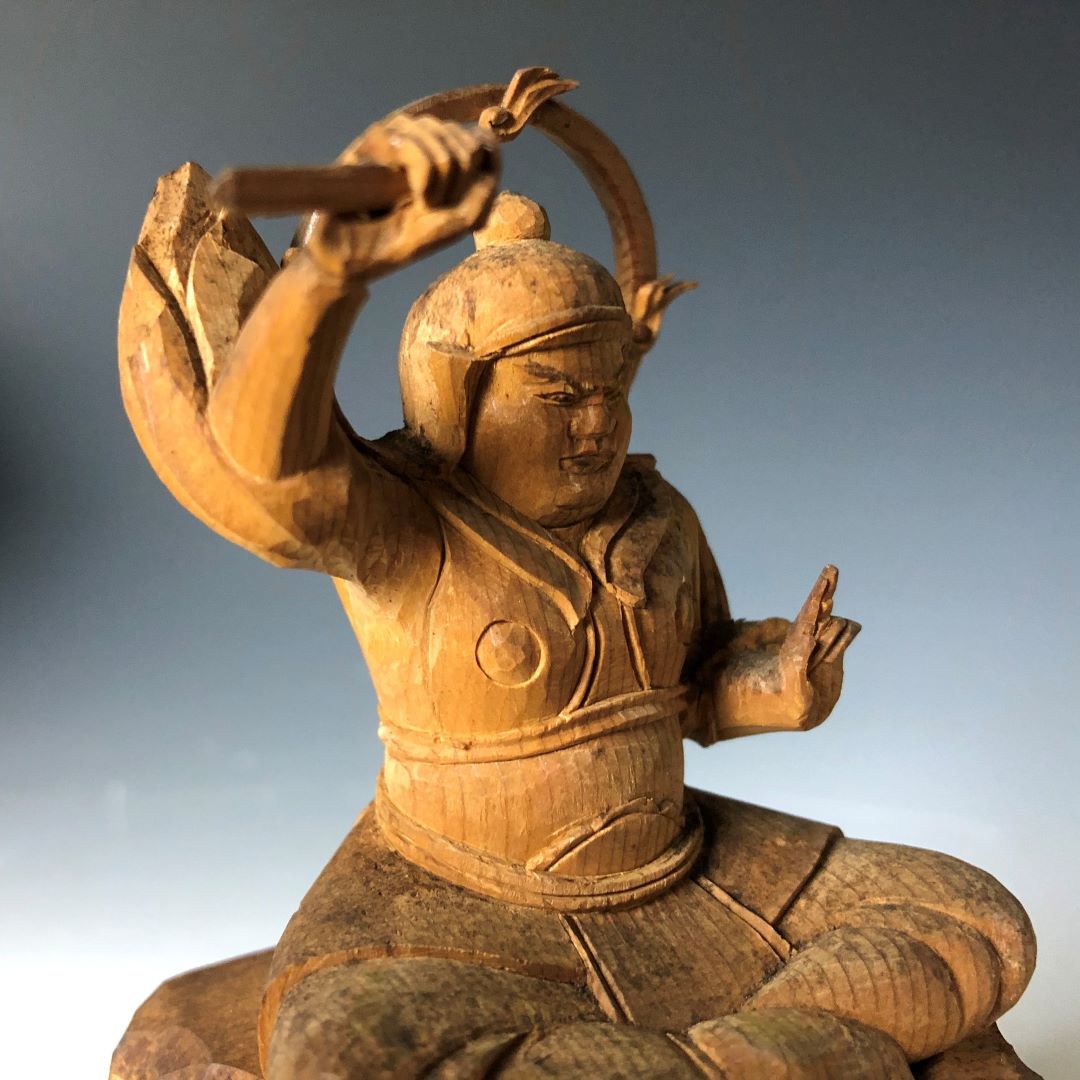 A wooden sculpture of a seated figure with a helmet, pointing upwards, holding a curved object, showcasing detailed traditional attire and expressive craftsmanship.
