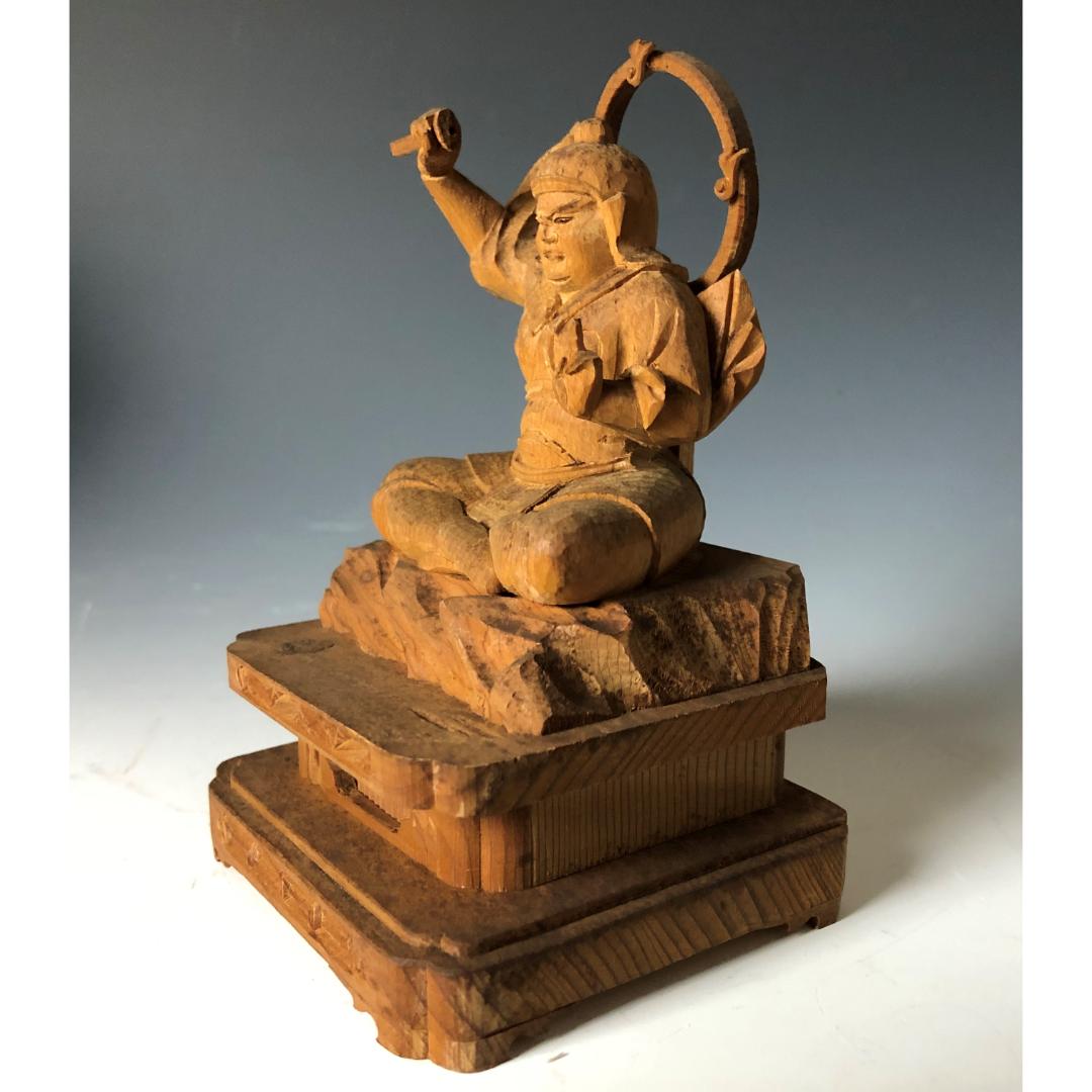 A wooden sculpture of a joyful figure seated cross-legged, with one arm raised and holding a ring, on a multi-tiered wooden base. The figure is carved with visible smiles and intricate detailing on the clothing and base.
