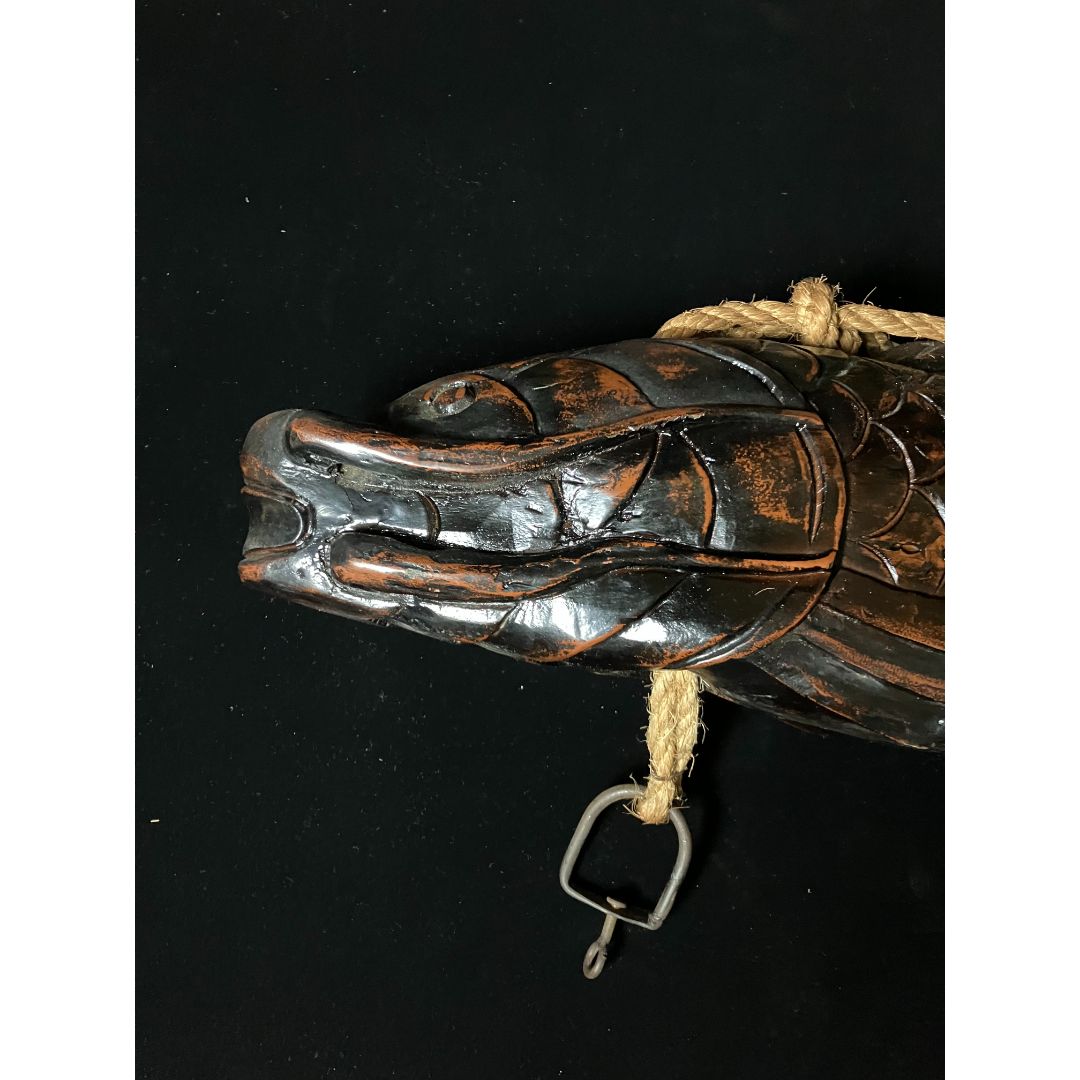 A carved wooden fish head is being used as a pot carrier. The fish head has intricate carvings and a hook, with a thick rope passing through its head. It is placed on a black cloth, showcasing its unique features.