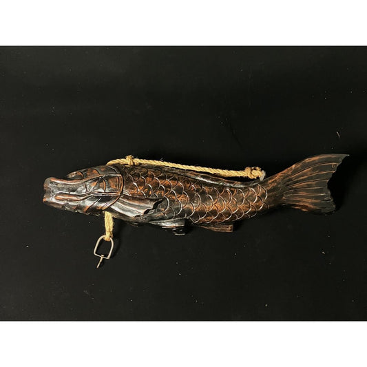A wooden fish with intricate carvings is being used as a pot carrier. The fish has a hook and a thick rope passing through its body. It is placed on a black cloth, highlighting its unique features.