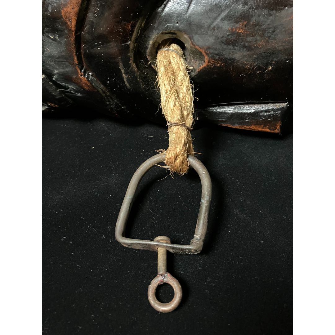 An iron device with a hook is attached to a thick rope, which is hanging from a jizaikagi. The jizaikagi is a traditional Japanese fireplace crane used to suspend pots over an open fire. The device and rope are used to adjust the height of the pot over the fire.